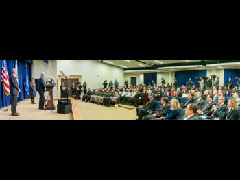 A wide-angle image of a man giving a speech from a podium on a stage to a crowd of seated people at the Medal of Valor award ceremony.